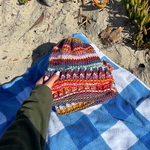 May include: A colorful striped knitted hat with a variety of colors including red, orange, yellow, blue, purple, and white. The hat is lying on a blue and white checkered towel.