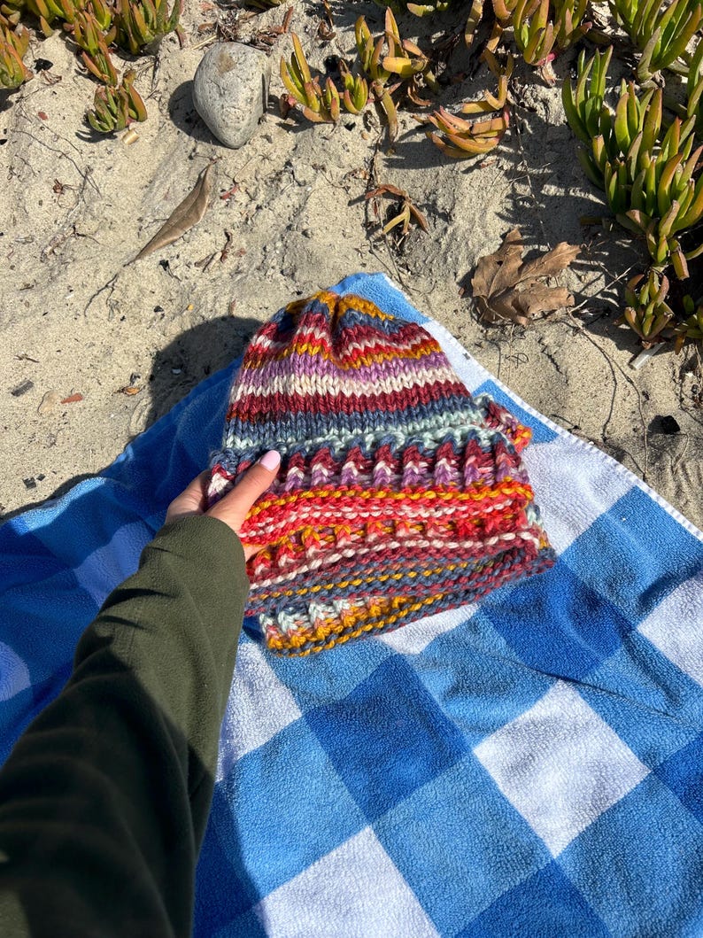 May include: A colorful striped knitted hat with a variety of colors including red, orange, yellow, blue, purple, and white. The hat is lying on a blue and white checkered towel.