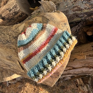 May include: A hand-knitted beanie hat with a ribbed brim and colorful stripes. The hat features a mix of colors including cream, blue, pink, and brown. The brim is a textured pattern in shades of blue, green, and yellow.