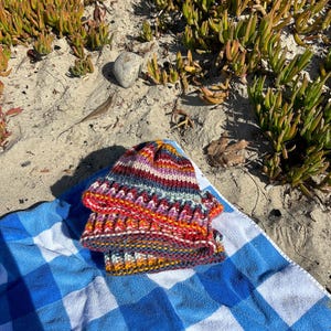 May include: A colorful striped knitted hat with a variety of colors including red, orange, yellow, blue, and purple. The hat is folded in half and placed on a blue and white checkered towel.