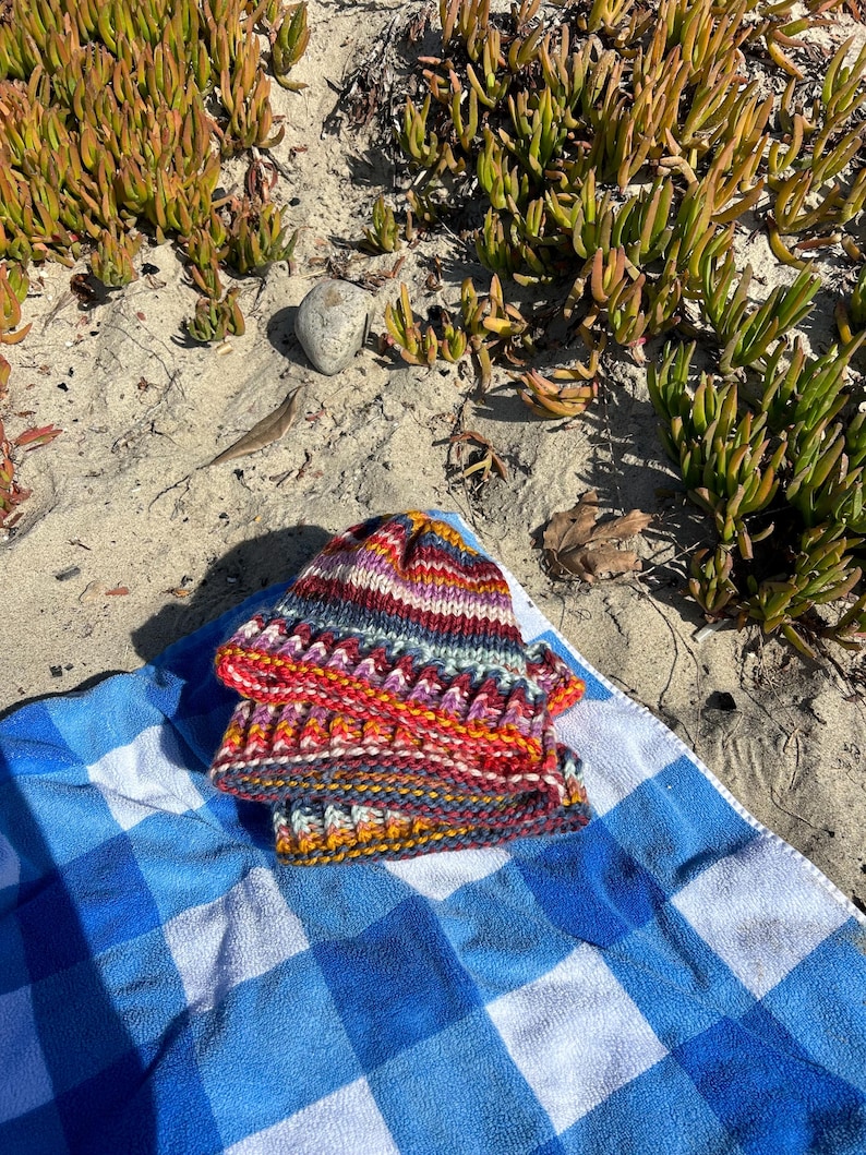 May include: A colorful striped knitted hat with a variety of colors including red, orange, yellow, blue, and purple. The hat is folded in half and placed on a blue and white checkered towel.