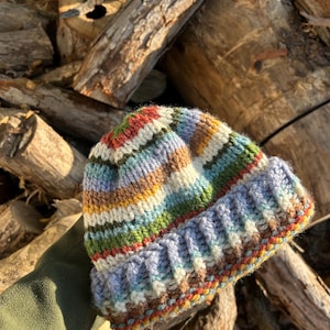 May include: A hand-knitted beanie featuring a multi-colored striped pattern in shades of blue, green, yellow, brown, and cream. The hat has a thick, ribbed folded brim and a textured knit construction, showcasing a variety of yarn colors throughout.