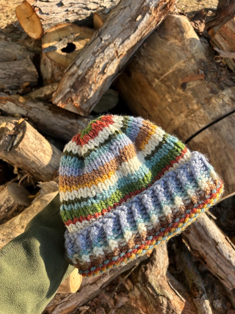 May include: A hand-knitted beanie featuring a multi-colored striped pattern in shades of blue, green, yellow, brown, and cream. The hat has a thick, ribbed folded brim and a textured knit construction, showcasing a variety of yarn colors throughout.