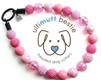 May include: Pink beaded dog collar with a black clasp and a small charm. The collar has a pink and white iridescent finish. The text "ultimutt bestie beaded dog collars" is printed on a white background.