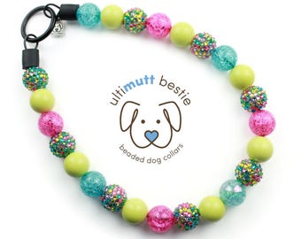 Puede incluir: Un collar de perro con cuentas de colores con cuentas rosas, verdes y azules. El collar presenta el texto "ultimutt bestie" y un gráfico de perro. Tiene un cierre de metal negro.