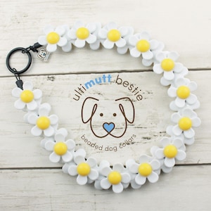 May include: A dog collar featuring a series of white daisy-shaped beads with yellow centers. The collar includes a black metal ring and a small tag with a dog logo. The text "ultimutt bestie beaded dog collars" is printed in the center.