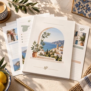 May include: A stack of white paper planners featuring a Mediterranean coastal scene printed on the cover, showing a stone archway, a potted plant, white buildings, and a blue sea. The pages include lined sections, a map of Spain, and leaf icons.
