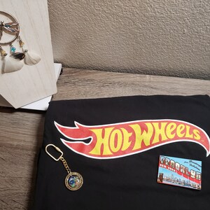 May include: Black t-shirt with the Hot Wheels logo in red and yellow. A gold keychain with a globe charm and a postcard with the text "Brooklyn" are on the shirt.