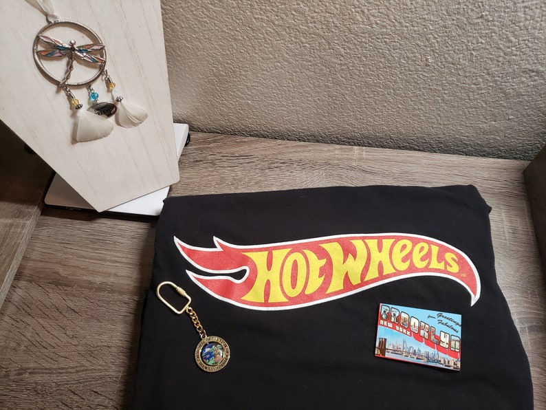 May include: Black t-shirt with the Hot Wheels logo in red and yellow. A gold keychain with a globe charm and a postcard with the text "Brooklyn" are on the shirt.