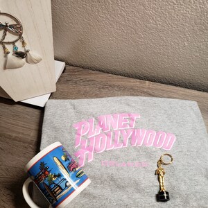 May include: A gray t-shirt with the pink text "Planet Hollywood Orlando" printed on the front. A gold-colored keychain with a small statue of a person is on the shirt. A blue and white mug with a beach scene is in the foreground.