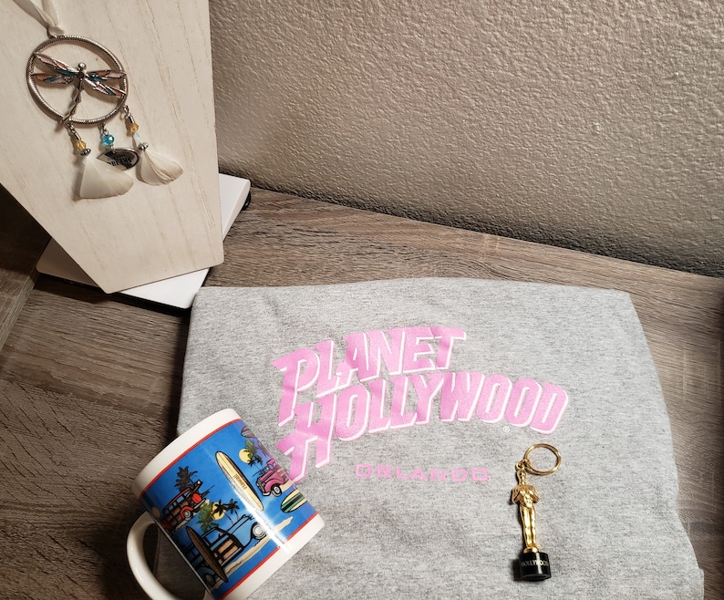 May include: A gray t-shirt with the pink text "Planet Hollywood Orlando" printed on the front. A gold-colored keychain with a small statue of a person is on the shirt. A blue and white mug with a beach scene is in the foreground.