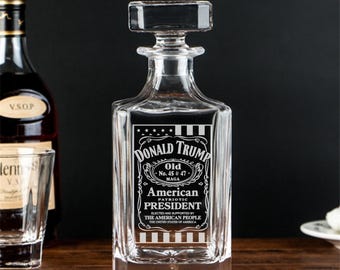 May include: A clear glass decanter with a square base and a square stopper. The decanter is engraved with an American flag design and the text "DONALD TRUMP Old No. 45 & 47 MAGA American PATRIOTIC PRESIDENT ELECTED AND SUPPORTED BY THE AMERICAN PEOPLE THE UNITED STATES OF AMERICA".