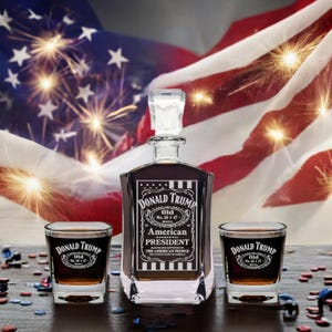 May include: A crystal decanter and two shot glasses with an engraved design featuring the text "Donald Trump, Old No. 45 & 47, MAGA, American Patriotic President, Elected and Supported by the American People". The decanter and shot glasses are on a wooden surface with a red, white, and blue American flag in the background.