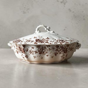 May include: A white ceramic tureen with a scalloped edge and a matching lid featuring a decorative handle. The exterior is adorned with a detailed brown floral and vine pattern. The vessel includes two small side handles for lifting and carrying.