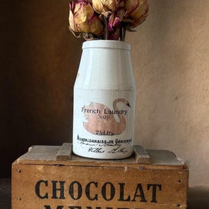May include: A white ceramic jar labeled French Laundry Soap with a swan graphic and 2 1/2 Ltr text, containing dried roses, resting on a wooden crate with the black text CHOCOLAT MENIER printed on the front side of the rectangular box.