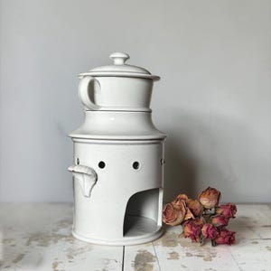 May include: A white ceramic fondue pot with a matching lid and a side handle. The base features a large arched opening for a heat source and a series of small circular ventilation holes around the upper rim. A cluster of dried pink roses sits nearby.