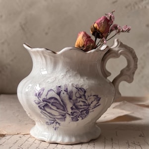 May include: A white ceramic pitcher with a purple floral design and a decorative handle. The pitcher has a cracked glaze and is filled with dried roses. The pitcher is sitting on a piece of paper with handwritten text.