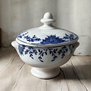 May include: A white ceramic serving bowl with a lid, featuring a blue floral pattern. The bowl has a pedestal base and small handles. The lid is topped with a small knob. The bowl is likely vintage or antique.