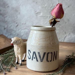 May include: A cream-colored ceramic jar with a crackled glaze finish and the word SAVON printed in black letters. A single dried pink rosebud is placed inside the jar, and a small white sheep figurine stands next to it on a wooden surface.