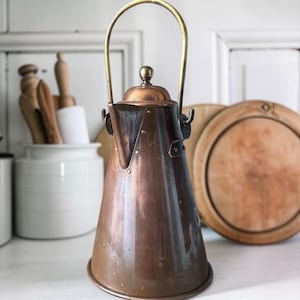 May include: A tall, conical copper kettle with a polished brass swing handle and a matching copper lid featuring a small spherical knob. The body of the kettle has a smooth, metallic finish with visible rivets securing the spout and handle mounts.