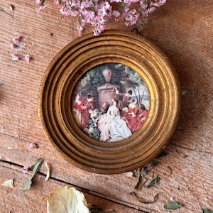 May include: A small, round, antique gold-toned picture frame with a detailed painting of people in a garden setting. The frame has a ridged design. Pink flowers and dried petals surround the frame on a wooden surface.