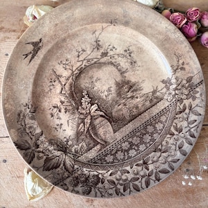 May include: A vintage decorative plate with a sepia-toned transferware design. The plate features a detailed landscape scene with a bird, floral elements, and fruit motifs. The rim has a decorative border. The plate is set on a wooden surface.