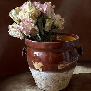 May include: A brown glazed ceramic pot with two side handles, featuring a textured, light-colored base and a smooth, dark upper section with decorative cream-colored markings. A bouquet of dried, pale pink and yellow roses sits inside the pot.