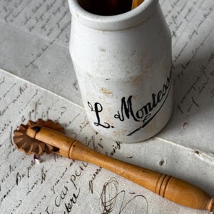 May include: An antique white ceramic jar with the text "L. Montessor" in black script. A wooden pastry cutter and wooden utensils are also visible. The items rest on a background of aged paper with handwritten script.