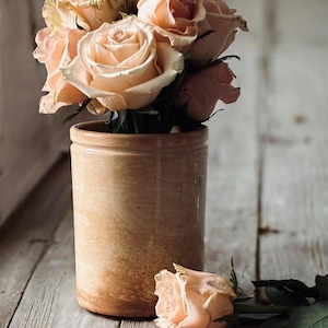 May include: A bouquet of peach-colored roses in a cylindrical, light brown ceramic vase. One rose lies on the wooden surface. The roses have multiple layers of petals, and the vase has a rustic appearance.