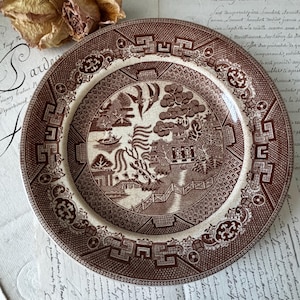 May include: A round ceramic plate featuring a brown and white transferware pattern. The central design depicts a landscape with a pagoda, a bridge, a boat, and two birds in flight. The wide rim is decorated with geometric and floral motifs.