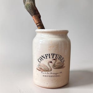 May include: A vintage ceramic jar with the word "CONFITURES" and two swans. The jar has a brush inside. The text "Rue de la Montagne 26. BRUXELLES." is below the swans. The jar is off-white.