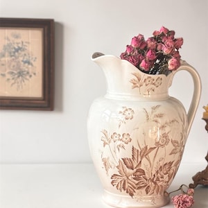 May include: Cream-colored ceramic pitcher with a floral design in brown tones. The pitcher is filled with dried pink flowers. A framed floral print is in the background, adding to the vintage aesthetic. The pitcher has a handle and a spout.