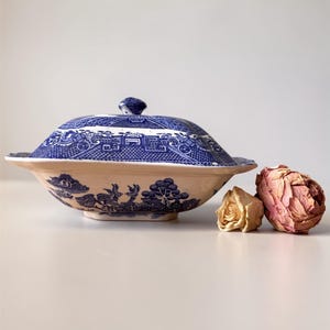 May include: A blue and white ceramic serving dish with a lid, featuring a classic Willow pattern design. The dish is oval-shaped with a lid and a small handle. Two dried roses are next to the dish.