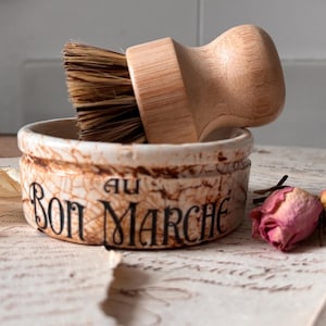 May include: A rustic, ceramic dish with the text "AU BON MARCHE" in black script, holding a wooden-handled brush with brown bristles. Dried rose buds and a piece of paper are in the foreground.