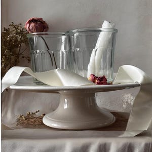 May include: A white ceramic cake stand with a round top and a pedestal base, holding two clear glass jars and white candles. A white ribbon and dried flowers are draped around the stand, creating a rustic, elegant display.