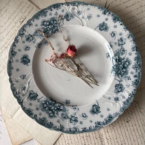 May include: A round ceramic plate with a scalloped edge, featuring a white base and a blue floral transferware pattern. A small cluster of dried pink and red flowers is placed in the center of the plate, which has embossed details.