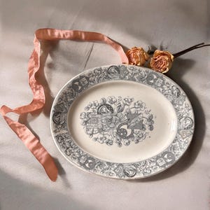 May include: An oval, antique-style serving platter with a detailed gray floral and geometric pattern around the rim and center. A peach-colored ribbon and dried roses are arranged near the platter. The platter is approximately 12 inches long.