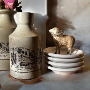 May include: An antique ceramic bottle with black text and a decorative label stands next to a stack of small white plates. A small sheep figurine with a silver garland is on top of the plates. Dried roses are in a similar bottle.