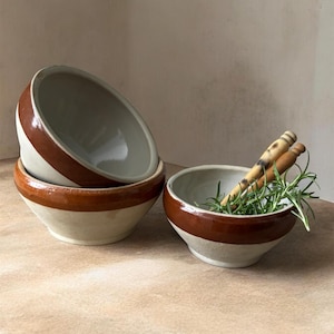 May include: A stack of three off-white ceramic bowls with a wide brown band around the rim. One bowl contains rosemary sprigs and two wooden stirrers. The bowls are on a light brown surface.