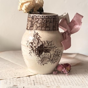 May include: A vintage ceramic pitcher with a floral design in brown and cream. The pitcher has a handle and is adorned with a pink ribbon. A dried rose and small pink flowers are placed inside and near the pitcher. The pitcher sits on aged paper.