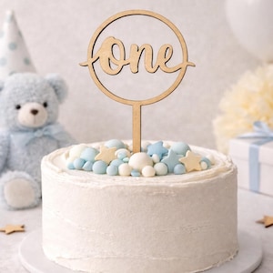Könnte beinhalten: Ein weißer Kuchen mit blauer und weißer Süßigkeiten- und sternförmiger Dekoration. Ein Kuchenaufsatz aus Holz mit dem Wort "one" befindet sich in der Mitte. Ein blauer Teddybär und eine Partyhütchen sind im Hintergrund.
