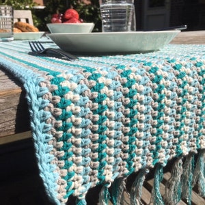 May include: A crocheted table runner with a blue, white, and teal geometric pattern. The runner has fringe on the ends.
