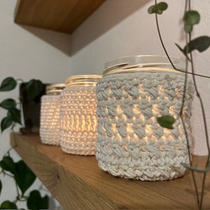 May include: Four glass jars with white crocheted covers, lit from within, creating a warm glow. The jars are arranged on a wooden shelf.