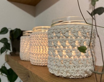 May include: Three clear glass jars are each encased in a thick, off-white crocheted cover with a textured, repeating stitch pattern. Each jar contains a warm, glowing light source that illuminates the interior, casting a soft light through the yarn.