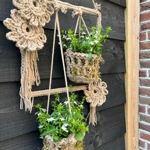 May include: Two macrame plant hangers with white flowers hanging from a black wooden wall. The plant hangers are made of natural rope and have a flower design at the top.