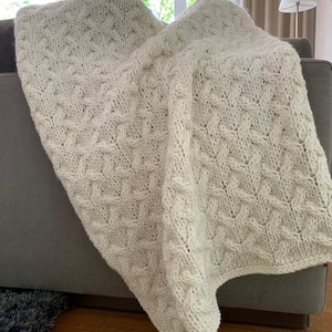 May include: A white knitted throw blanket with a cable knit pattern. The blanket is draped over a gray couch.