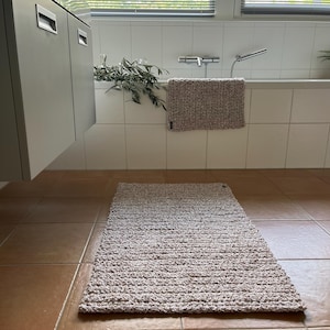 Puede incluir: Una alfombra de baño de color marrón claro con un diseño texturizado y tejido. La alfombra está colocada en un suelo de baldosas en un baño.