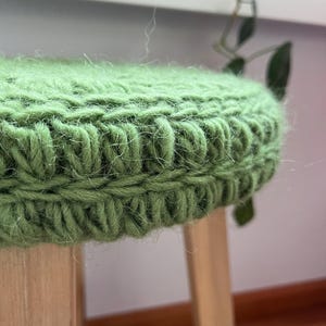 May include: A close-up of a green knitted stool cushion. The cushion is made of a soft, fuzzy yarn and has a simple, textured design.