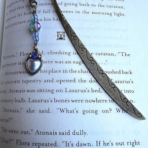 May include: A silver bookmark with an ornate design, featuring a small hourglass charm and a blue and white glass bead. The bookmark is placed on a page of a book with text about a caravan and a unicorn tapestry.