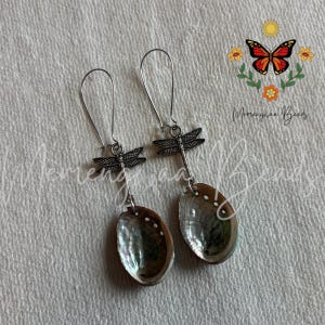 May include: A pair of dangle earrings featuring silver-toned dragonfly charms attached to oval-shaped iridescent abalone shell pendants. The earrings include long, thin silver-toned ear wires for hanging. The image includes a butterfly logo and text.